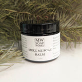 A 2 ounce jar of Sore Muscle Balm from MW Soapworks sits in greenery against a wood background.