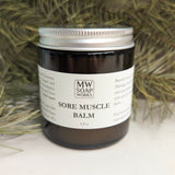 A 4 ounce jar of Sore Muscle Balm from MW Soapworks sits in front of greenery and a woody background.