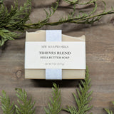 A tan bar of Thieves Blend Shea Butter Soap from MW Soapworks sits on a wooden background with cedar greenery.