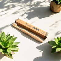 A 100% plastic free toothbrush for kids sits on a dappled sunlit background surrounded by plants. It sits next to its packaging box, a kraft box indicating that it is a bamboo toothbrush with natural bristles.