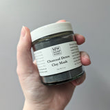 A hand holds up a jar of MW Soapworks Activated Charcoal Detox Clay Mask.