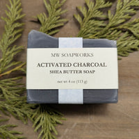A dark four ounce natural soap bar from MW Soapworks sits on a cedar branch. The label says it is an Activated Charcoal Shea Butter Soap.