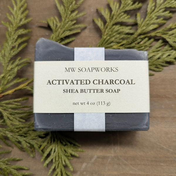 A dark four ounce natural soap bar from MW Soapworks sits on a cedar branch. The label says it is an Activated Charcoal Shea Butter Soap.