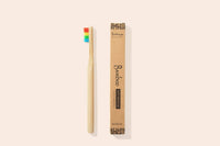 A bamboo toothbrush with rainbow bristles sits next to its box against a pale pink background. The box indicates it is a toothbrush with soft bristles.