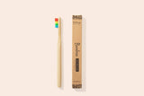 A bamboo toothbrush with rainbow bristles sits next to its box against a pale pink background. The box indicates it is a toothbrush with soft bristles.
