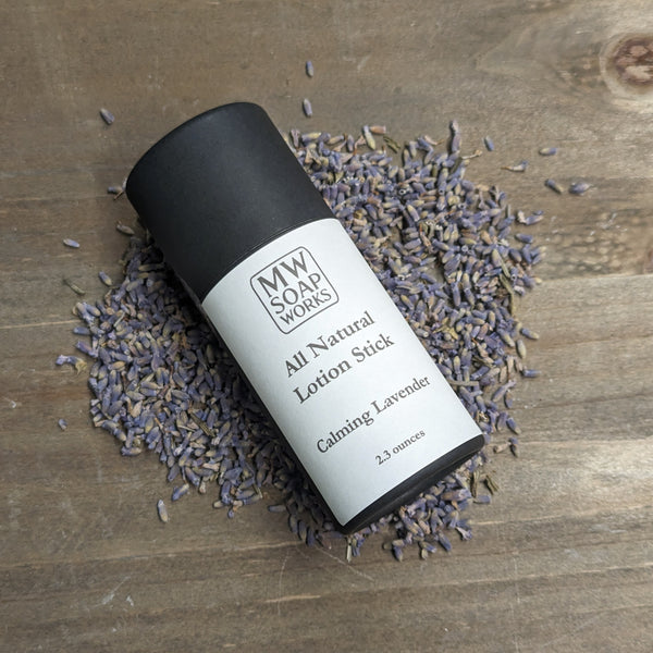 A black paperboard tube of Calming Lavender All Natural Lotion Stick from MW Soapworks sits surrounded by lavender buds on a wooden background.