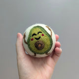 Hand holding a plush avocado wool dryer ball with a smiling face against a gray background