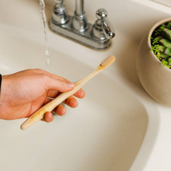 A hand holds a bamboo toothbrush in front of a white sink with the water running.