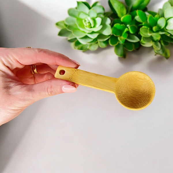 A hand holds a shallow all natural bamboo spoon in front of plants and a white background.