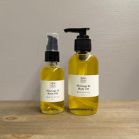 Two clear glass bottles of MW Soapworks Massage and Body Oil sit on a wooden shelf. The bottles contain 2 ounces and 4 ounces of Black Mission Fig Massage and Body Oil and have pump lids.