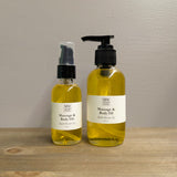 Two clear glass bottles of MW Soapworks Massage and Body Oil sit on a wooden shelf. The bottles contain 2 ounces and 4 ounces of Black Mission Fig Massage and Body Oil and have pump lids.