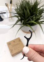 A hand holds a biodegradable dental floss pick in a bathroom.