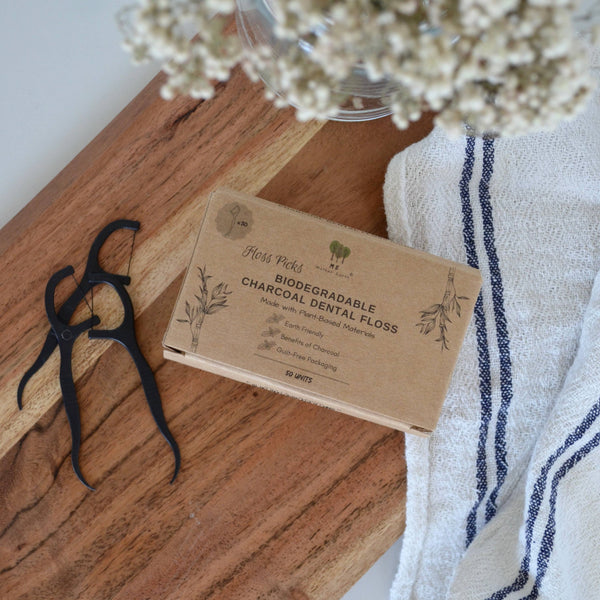 Biodegradable Charcoal Dental Floss Picks sit on a wooden background with a tea towel and flowers.