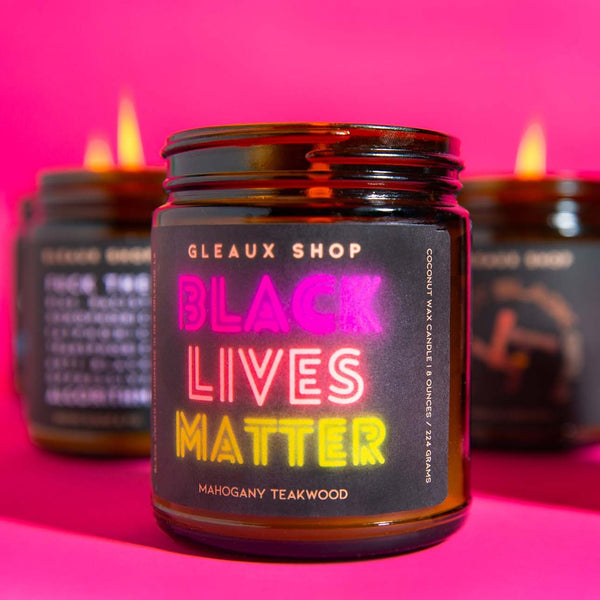 A candle packaged in an amber jar sits in front of a pink background. The label reads "Black Lives Matter"