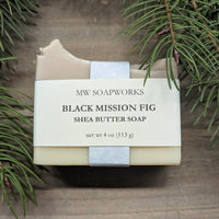 A 4 ounce bar of MW Soapworks Black Mission Fig Shea Butter Soap sits against greenery and a natural wood background.