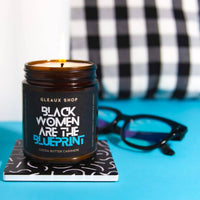 A candle packaged in an amber jar sits on a coaster on a blue table. The label reads "Black Women are the Blueprint"