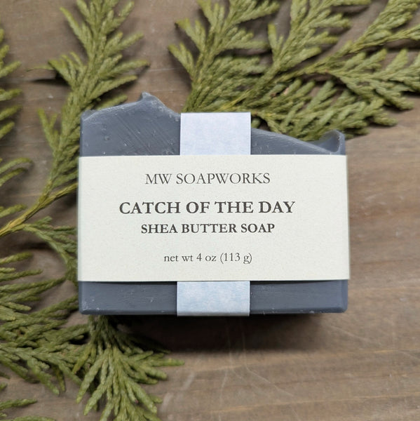 A dark gray soap sits on a natural background of wood and cedar branches. The label reads MW Soapworks Catch of the Day Shea Butter Soap net wt 4 oz (113 g)
