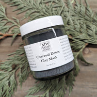 A glass jar of MW Soapworks Charcoal Detox Clay Mask sits on a cedar branch and a wood background.
