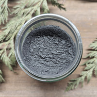 A jar of MW Soapworks Charcoal Detox Clay Mask sits open on a wooden background. The clay mixture within is a dark grey color.