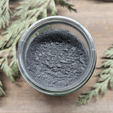 A jar of MW Soapworks Charcoal Detox Clay Mask sits open on a wooden background. The clay mixture within is a dark grey color.