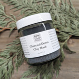A glass jar of MW Soapworks Charcoal Detox Clay Mask sits on a cedar branch and a wood background.