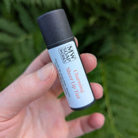 A hand holds up a black compostable paper tube of Charming Sheer Lip Tint from MW Soapworks, which is a light pink/nude color.