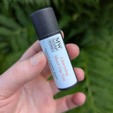 A hand holds up a black compostable paper tube of Charming Sheer Lip Tint from MW Soapworks, which is a light pink/nude color.