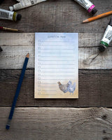 Featuring the words "scratch pad" at the top, a notepad sits against a wooden background surrounded by art supplies. The note pad features watercolor illustrations of cute chickens.