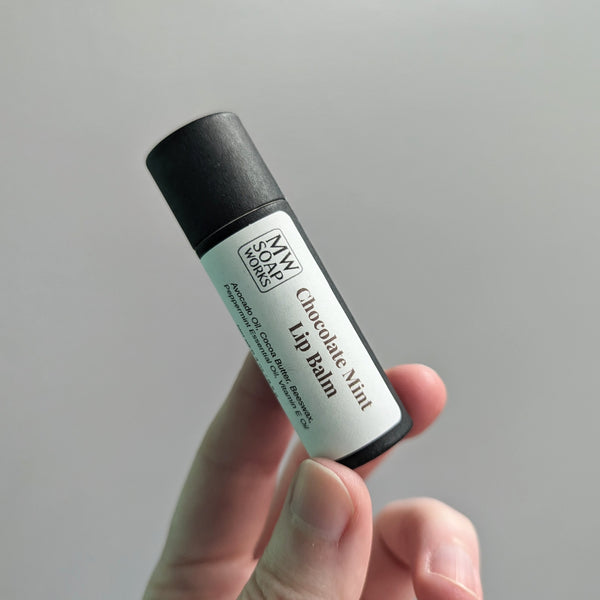 A hand holds up a lip balm packaged in a black compostable paper tube. The label of the lip balm has the MW Soapworks logo and the words Chocolate Mint Lip Balm with the ingredients below.