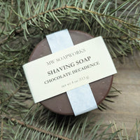 A round bar of MW Soapworks Chocolate Decadence Shaving Soap sits among branches from a PNW forest.