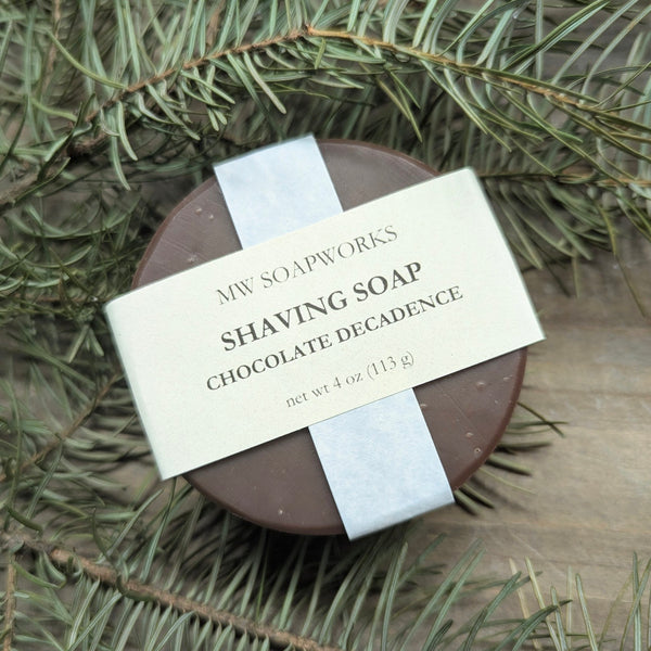 A round bar of MW Soapworks Chocolate Decadence Shaving Soap sits among branches from a PNW forest.