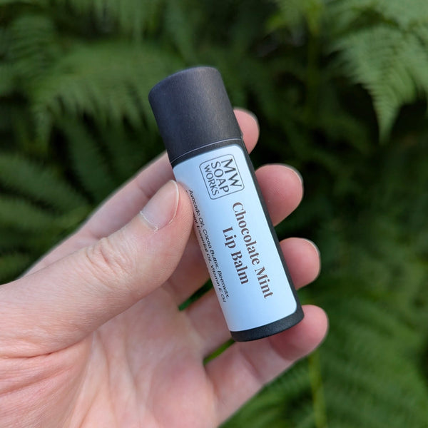 A hand holds up a black paper compostable tube of MW Soapworks Chocolate Mint Lip Balm in front of lush green Pacific Northwest ferns.