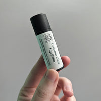 A hand holds up a lip balm packaged in a black compostable paper tube. The label of the lip balm has the MW Soapworks logo and the words Coconut Lime Lip Balm with the ingredients below.