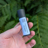 A hand holds up a black eco-friendly tube of Coconut Lime Lip Balm from MW Soapworks. There are lush green Pacific NW ferns in the background.