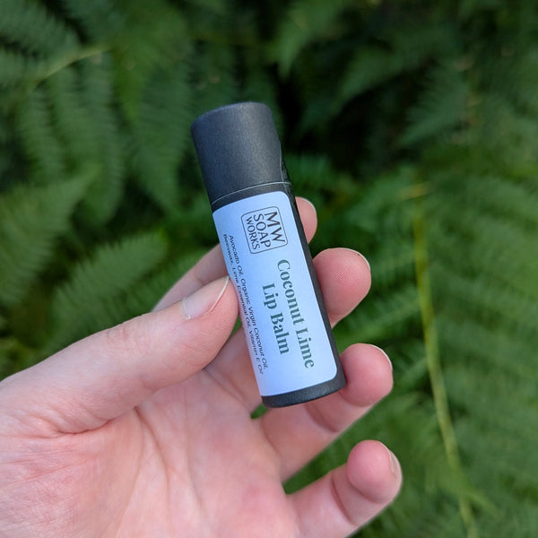 A hand holds up a black eco-friendly tube of Coconut Lime Lip Balm from MW Soapworks. There are lush green Pacific NW ferns in the background.