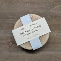 A round MW Soapworks bar soap sits against a natural wood background. It has a paper label indicating it is a 4 ounce bar of Coconut Oatmeal Shea Butter Soap.