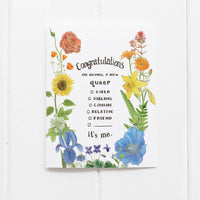 A greeting card with watercolor illustrations of a rainbow of colorful flowers surrounds a coming out message. The text reads "Congratulations on having a new queer child/sibling/cousin/relative/friend. It's Me."