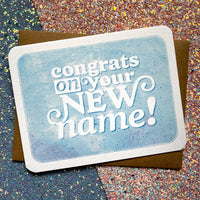 A blue greeting card has big text that reads "congrats on your new name!"