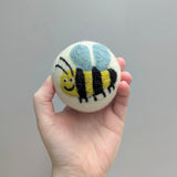 Hand holding a plush wool dryer ball with a cute bumblebee design against a gray background