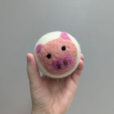 Hand holding a plush white wool dryer ball with a cute felted pig design against a gray background