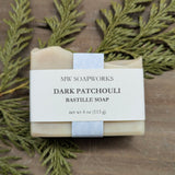 A 4 ounce bar of MW Soapworks Dark Patchouli Bastille Soap sits in front of a cedar branch against a wooden background.