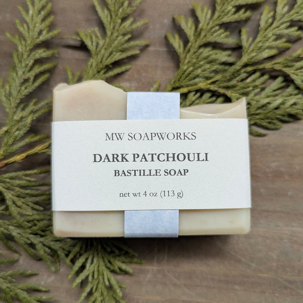 A 4 ounce bar of MW Soapworks Dark Patchouli Bastille Soap sits in front of a cedar branch against a wooden background.