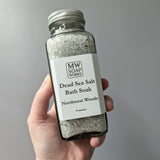 A hand holds up an 8 ounce square bottle of MW Soapworks Dead Sea Salt Bath Soak. The scent for the salt is Northwest Woods.