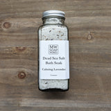 A clear glass bottle of MW Soapworks Dead Sea Salt Bath Soak sits on a wooden background. The label shows it is Calming Lavender scent.