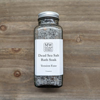 A glass bottle of MW Soapworks Tension Ease Dead Sea Salt Bath Soak sits against a natural wood background.