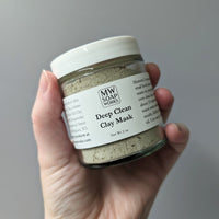A hand holds up a two ounce glass jar of MW Soapworks Deep Clean Clay Mask.