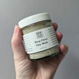 A hand holds up a two ounce glass jar of MW Soapworks Deep Clean Clay Mask.