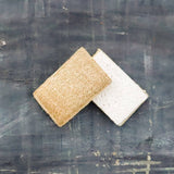 Two side of the same sponges, one brown loofah and one white wood pulp, on a textured dark surface with a pine cone.