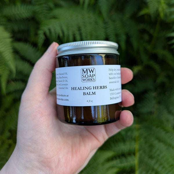 A hand holds a four ounce amber jar of MW Soapworks Healing Herbs Balm in front of PNW ferns.