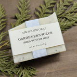 A four ounce bar of soap sits against a cedar branch. The label says it is a Gardener's Scrub Shea Butter Soap from MW Soapworks.